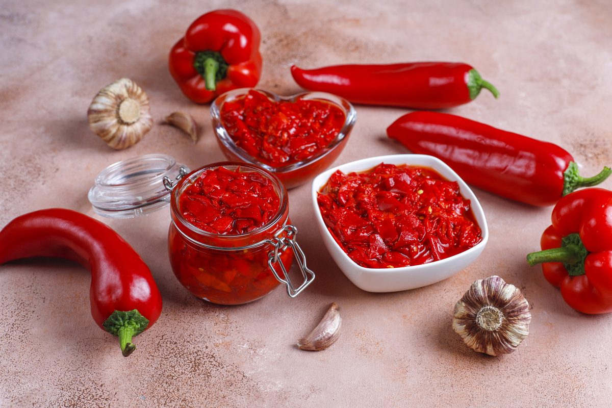 Сырая аджика из рубленого красного перца в банке и пиалах, рядом стручки перца и чеснок