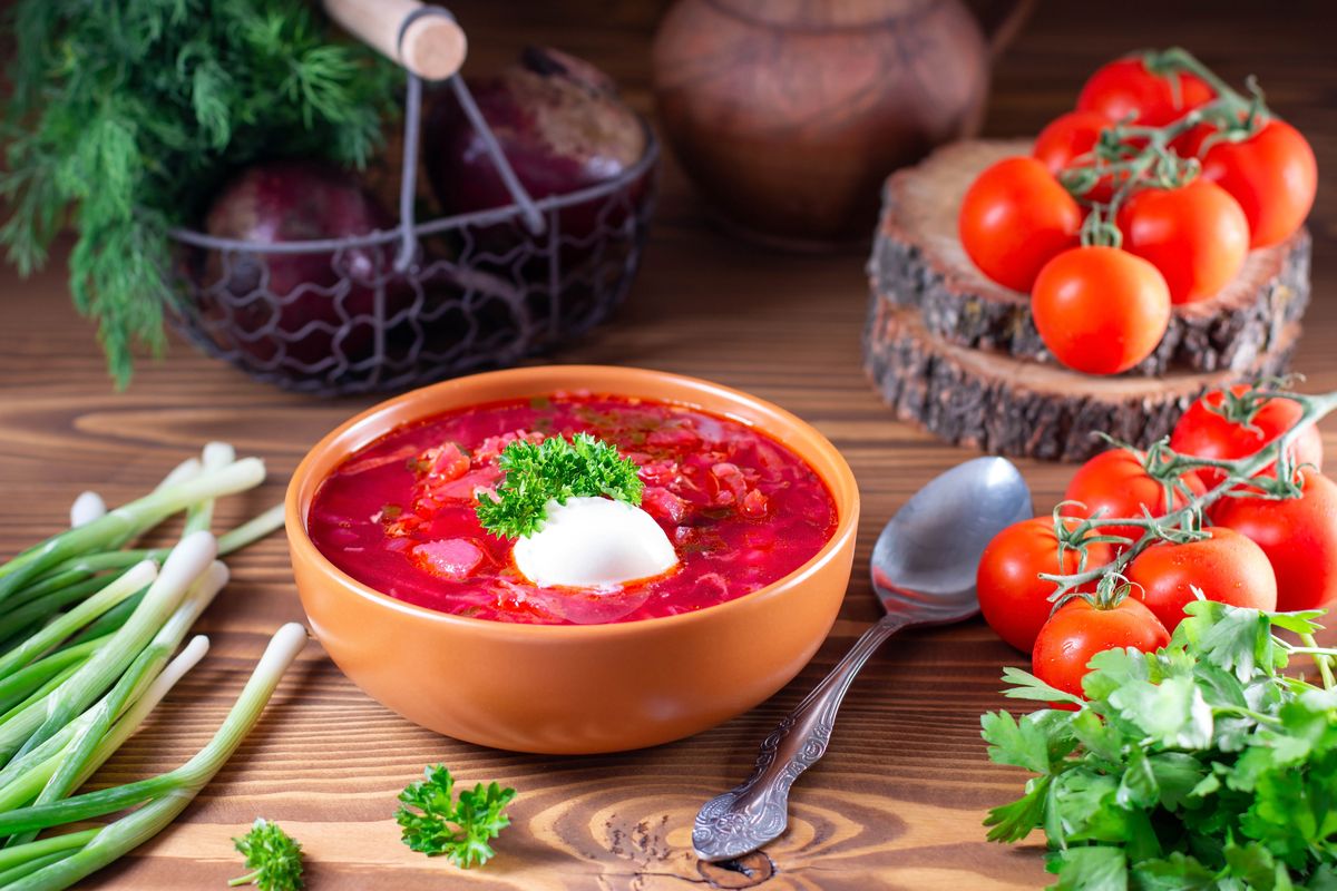 Борщ классический: как варить, на каком мясе, сколько настаивать Тарелка классического красного борща со сметаной и зеленью на деревянном столе, рядом помидоры и зелёный лук