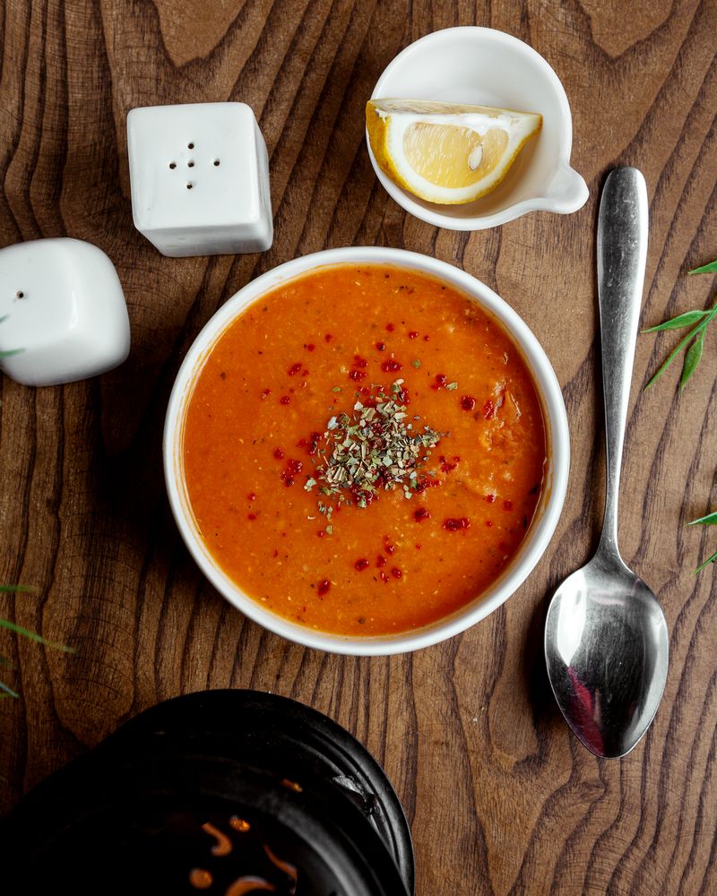 Гладкий оранжевый суп с сушёной мятой и хлопьями перца в масле, рядом долька лимона