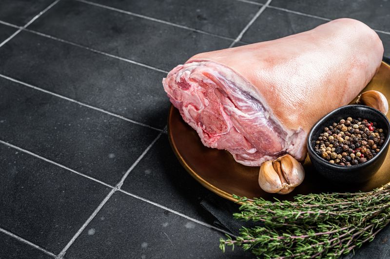 Сырая свиная рулька с кожей на тарелке рядом с перцем горошком и чесноком