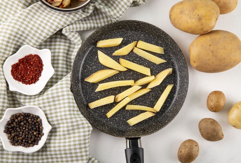 Сырые картофельные бруски в сковороде рядом с горошинами перца