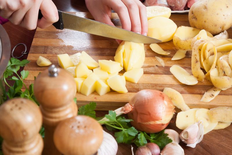 Очищенный картофель режут ножом на кубики на деревянной доске