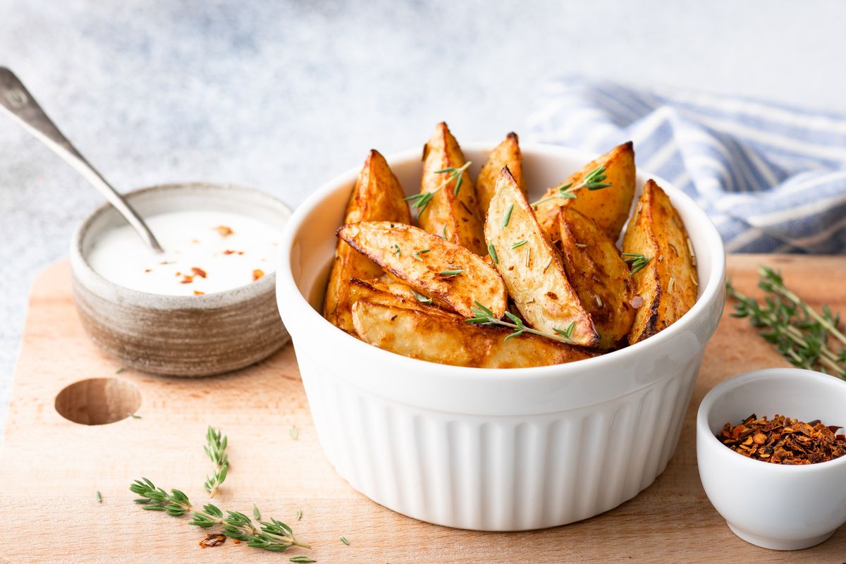 Картошка по-селянски в духовке Запечённые дольки картофеля в кожуре с паприкой и тимьяном в белой форме, рядом миска сметанного соуса