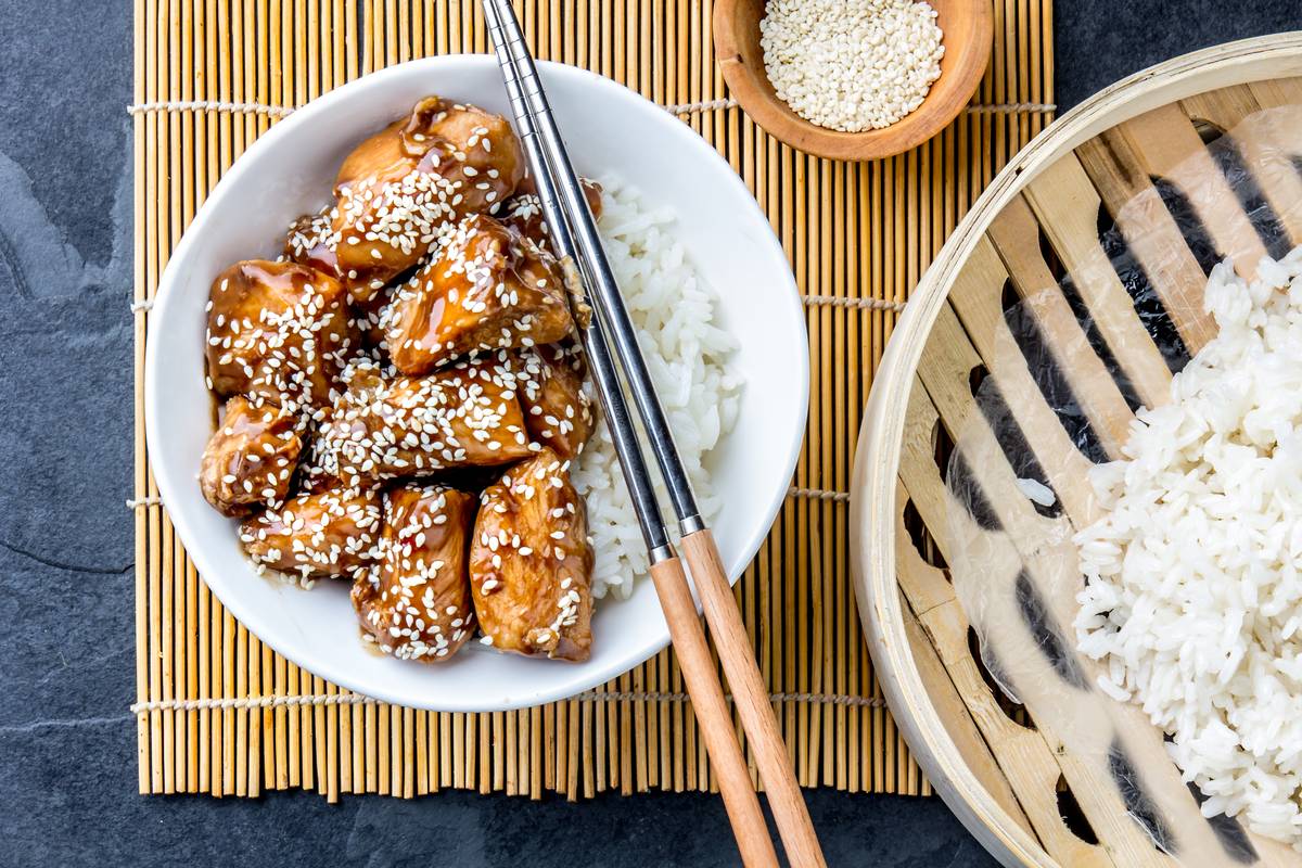 Кусочки курицы терияки в блестящей глазури с белым кунжутом, рядом отваренный рис и палочки