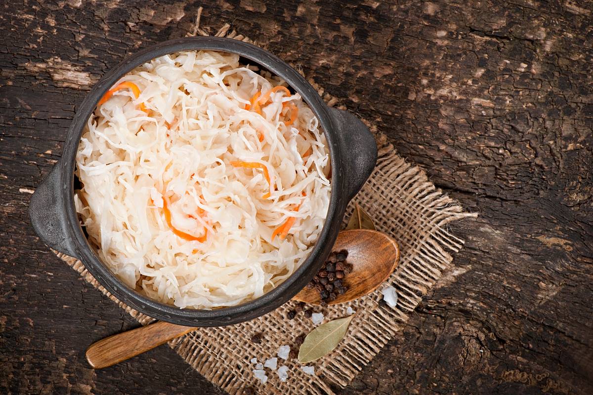 Квашеная капуста с морковью в чёрной керамической миске, рядом деревянная ложка с перцем горошком, лавровый лист и крупная соль