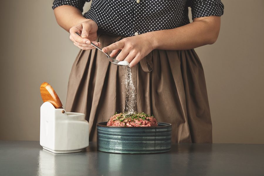 Соль сыплется из солонки в миску с мясным фаршем на кухонном столе