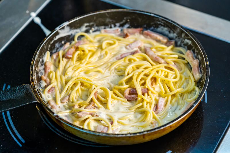 Спагетти со светлым сливочным соусом и кусочками бекона в сковороде на плите