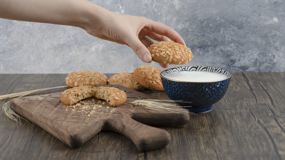 Овсяное печенье на деревянной доске рядом с миской молока, одно печенье в руке