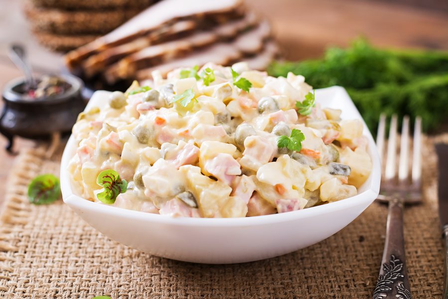 Готовый оливье в белой миске: видны кубики варёной колбасы, горошек и морковь