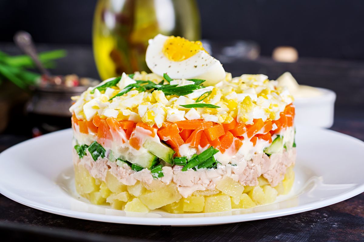Слоёный салат с печенью трески, картофелем, огурцом, морковью и яйцом на белой тарелке