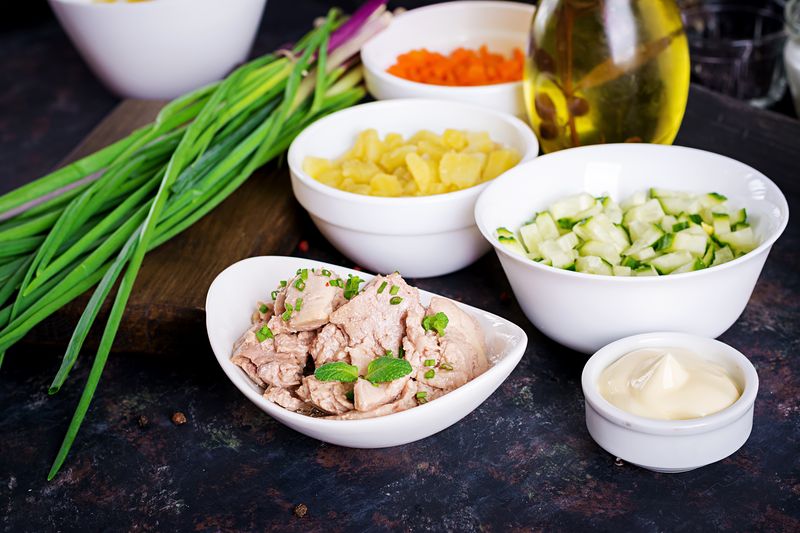 Печень трески кусочками в белой миске рядом с нарезанными огурцом и картофелем