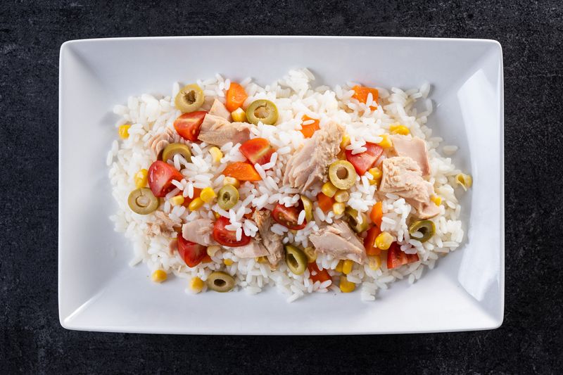 Рисовый салат с тунцом, помидорами, кукурузой и зелёными оливками на белом блюде