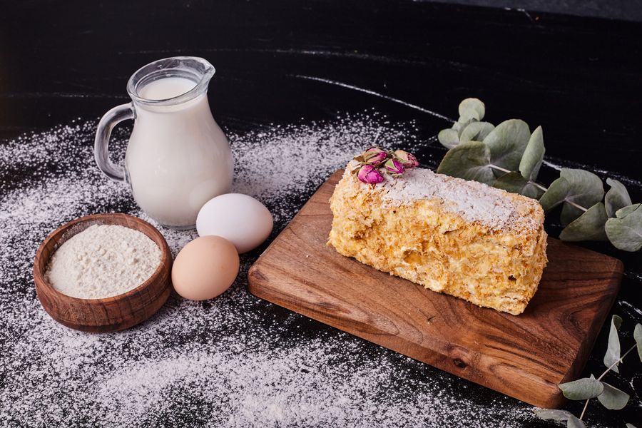 Кусок слоёного торта на деревянной доске рядом с кувшином молока, яйцами и миской муки