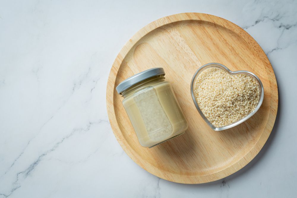 Банка світлої кунжутної пасти тахіні та мисочка очищеного насіння кунжуту на мармуровому столі