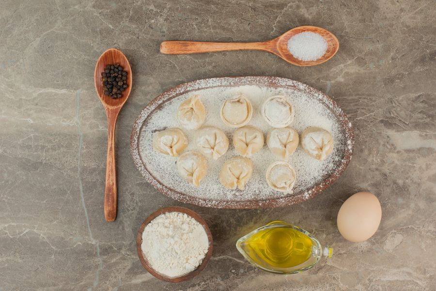 Сырые вареники на присыпанной мукой доске рядом с яйцом, маслом и перцем