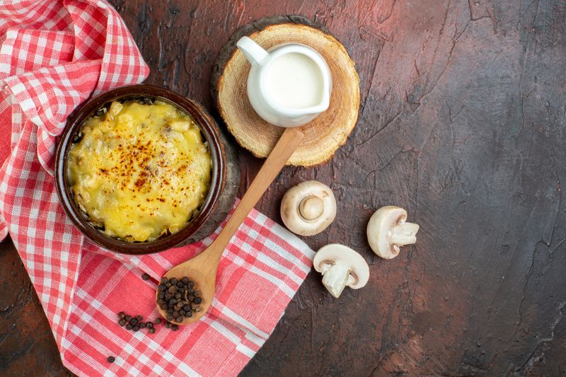Горшочек жульена рядом с молочником сливок, шампиньонами и ложкой чёрного перца